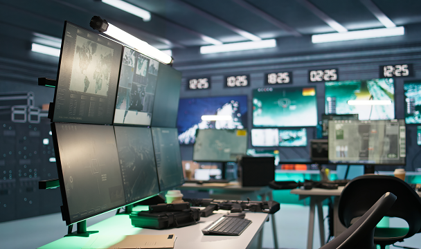 A modern control room workspace with multiple large monitors displaying maps, data dashboards, and video feeds, with a keyboard, mouse, and equipment on the desk in a high-tech, dimly lit environment.